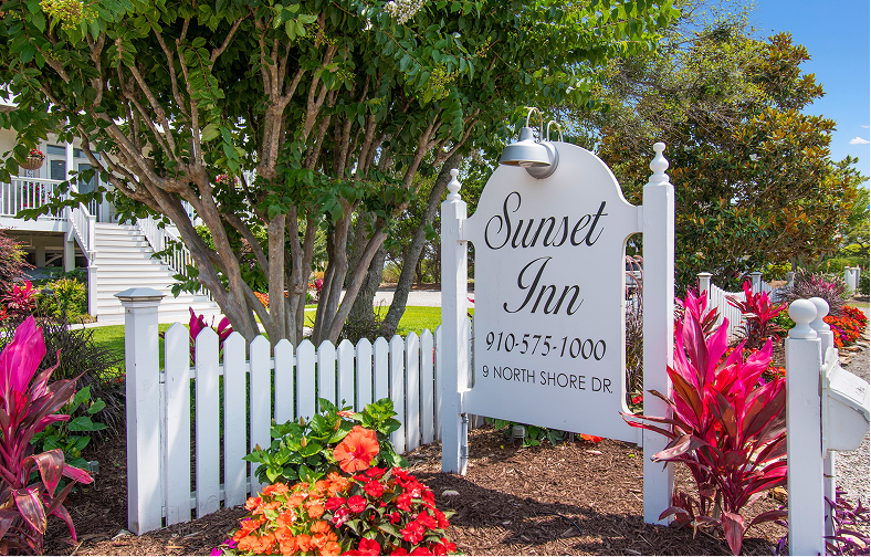 White picket fence and garden with colorful flowers surround a sign reading “Sunset Inn, 910-575-1000, 9 North Shore Dr.” Trees and a white building with stairs sit under a blue sky at this charming Sunset Beach NC bed and breakfast.