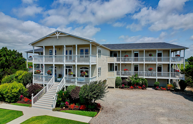 A large, light-colored, two-story Sunset Beach NC bed and breakfast with wraparound balconies, white railings, and potted flowers on each level. The property is surrounded by lush landscaping and sits under a partly cloudy sky.