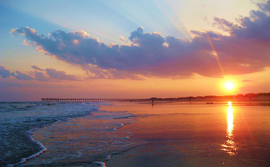 A vibrant Sunset Beach, NC sunset over gentle waves and a long pier; colorful clouds glow as the sun casts warm reflections on sand near a charming bed and breakfast.