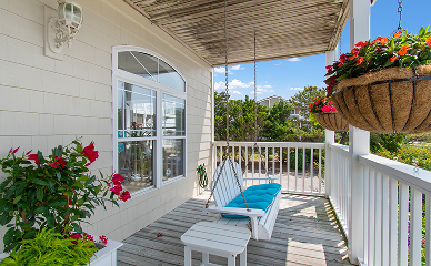 A porch at a Sunset Beach NC bed and breakfast features a white swing bench with a turquoise cushion, hanging basket of red flowers, potted plant, white railing, and wall lamp; trees and blue sky complete the peaceful scene.