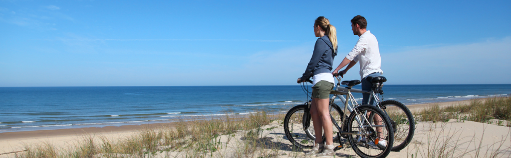 A man and a woman with bicycles stand on sandy dunes, looking out at the ocean under a clear blue sky—one of the scenic things to do in Sunset Beach NC as the beach and gentle waves stretch out below them.