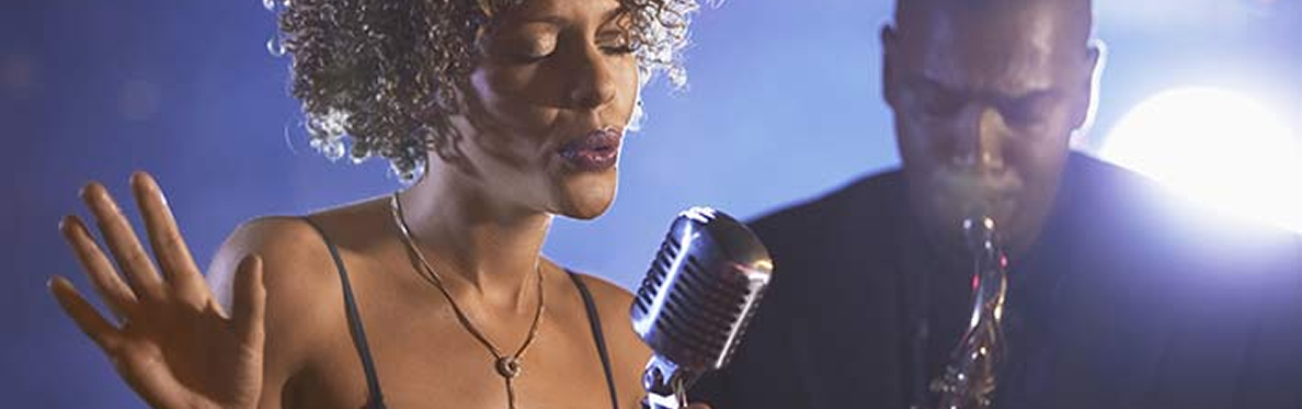 A woman with curly hair sings into a vintage microphone with her hand raised, while a man in the background plays a saxophone under dramatic stage lighting—a scene reminiscent of live music events among the things to do in Sunset Beach NC.