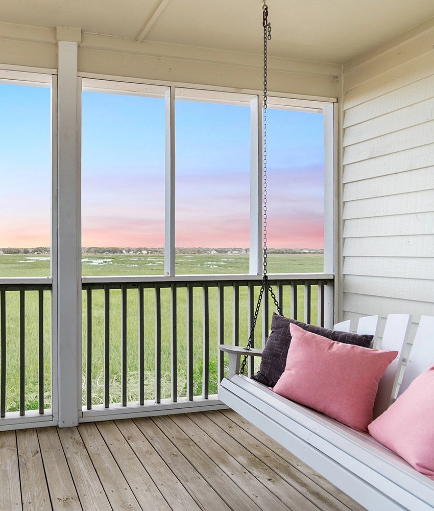 A cozy porch with a white swing bench topped with pink and gray pillows, overlooking a grassy landscape and water under a pastel Sunset Beach NC bed and breakfast sky.