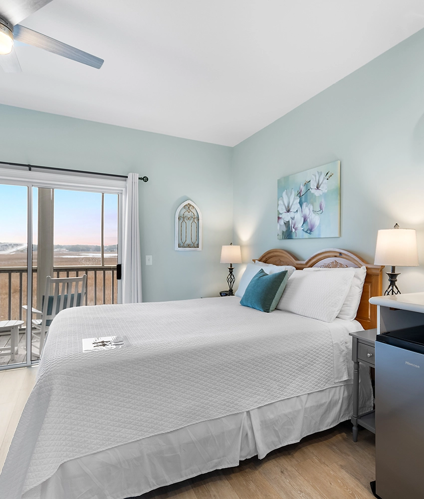A bright bedroom at this Sunset Beach NC bed and breakfast features a neatly made bed with white bedding, a teal accent pillow, bedside lamps, light blue walls, and a sliding glass door opening to a balcony with outdoor chairs and a scenic view.