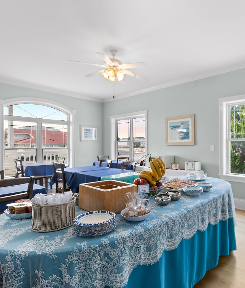 A breakfast buffet table with a lace tablecloth, plates, utensils, fruit, and pastries awaits guests at this Sunset Beach NC bed and breakfast in a bright dining room with blue walls, large windows, and several tables and chairs.