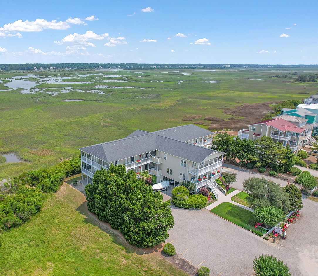 Aerial view of a large house with screened porches, surrounded by greenery and marshlands, with a gravel driveway—perfect for a Sunset Beach NC bed and breakfast—plus neighboring homes under a clear blue sky.