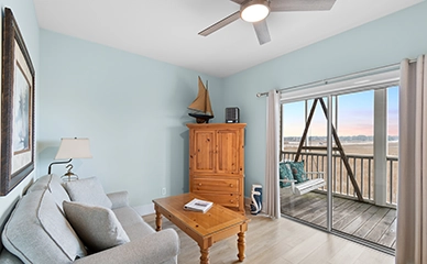 A cozy living room with a light blue wall, gray sofa, wooden coffee table, and cabinet. Sliding glass doors open to a balcony with a swing and scenic outdoor view at sunset—a perfect retreat inspired by a Sunset Beach NC bed and breakfast.