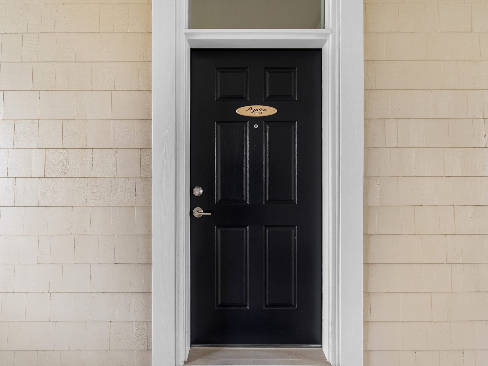 A black front door with six panels, a silver handle, and a small plaque above the peephole marks the entrance to the Azalea Room at Sunset Inn, set in a white frame against tan shingle siding.