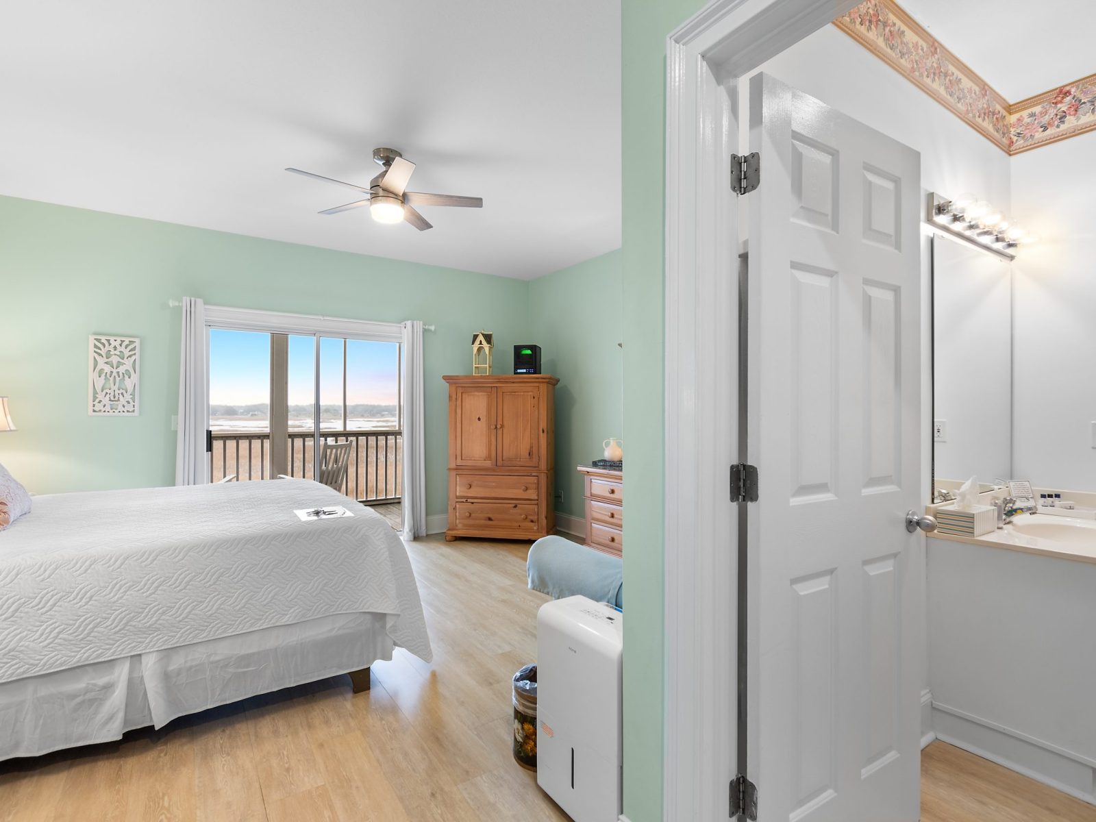 The Azalea Room at Sunset Inn features mint green walls, a large bed, wooden furniture, balcony doors with a scenic view, and an attached bathroom with a vanity and mirror.