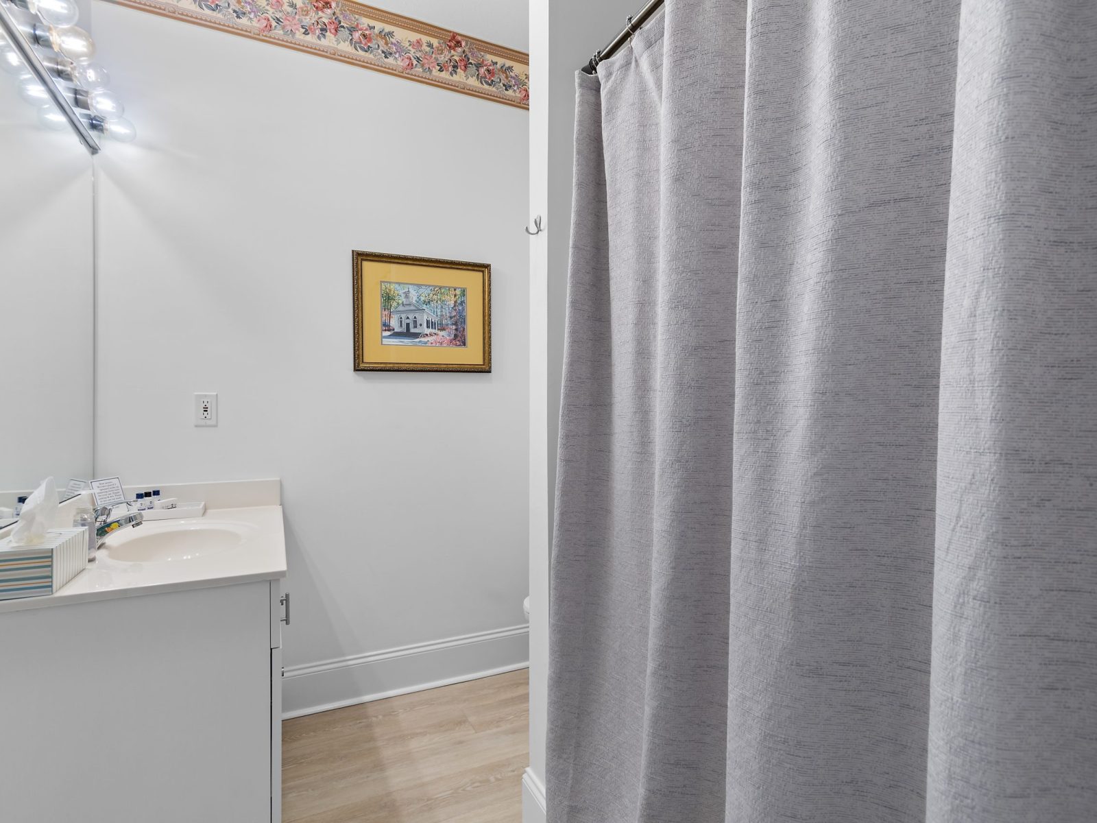 A bathroom in the Azalea Room at Sunset Inn features a white sink and countertop, mirror with lights, toiletries, framed artwork, floral wallpaper border, wood floor, and a partially closed gray shower curtain.