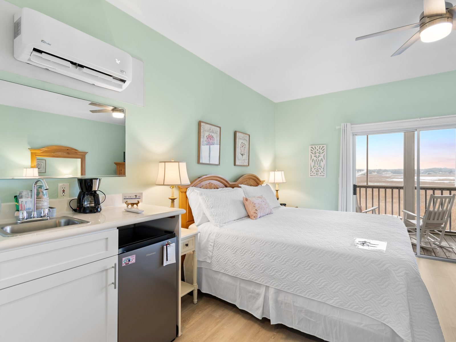 The Azalea Room at Sunset Inn features a tidy bedroom with a double bed, mint green walls, a kitchen area, and glass doors opening to a balcony with stunning beach and ocean views.