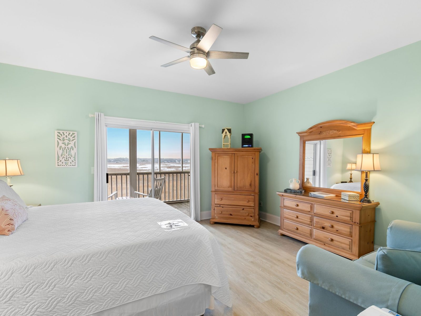 The Azalea Room at Sunset Inn features light green walls, a spacious bed with white bedding, wood furniture, a ceiling fan, and a sliding glass door opening to a balcony with an ocean view.