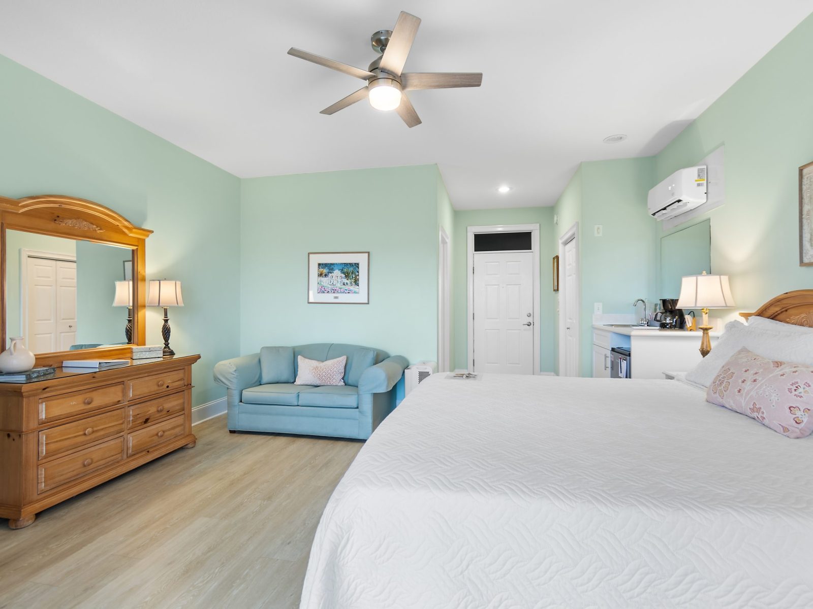 The Azalea Room at Sunset Inn features mint green walls, a large bed with white bedding, a wooden dresser with mirror, light blue loveseat, ceiling fan, and a kitchenette area with coffee maker for your comfort.