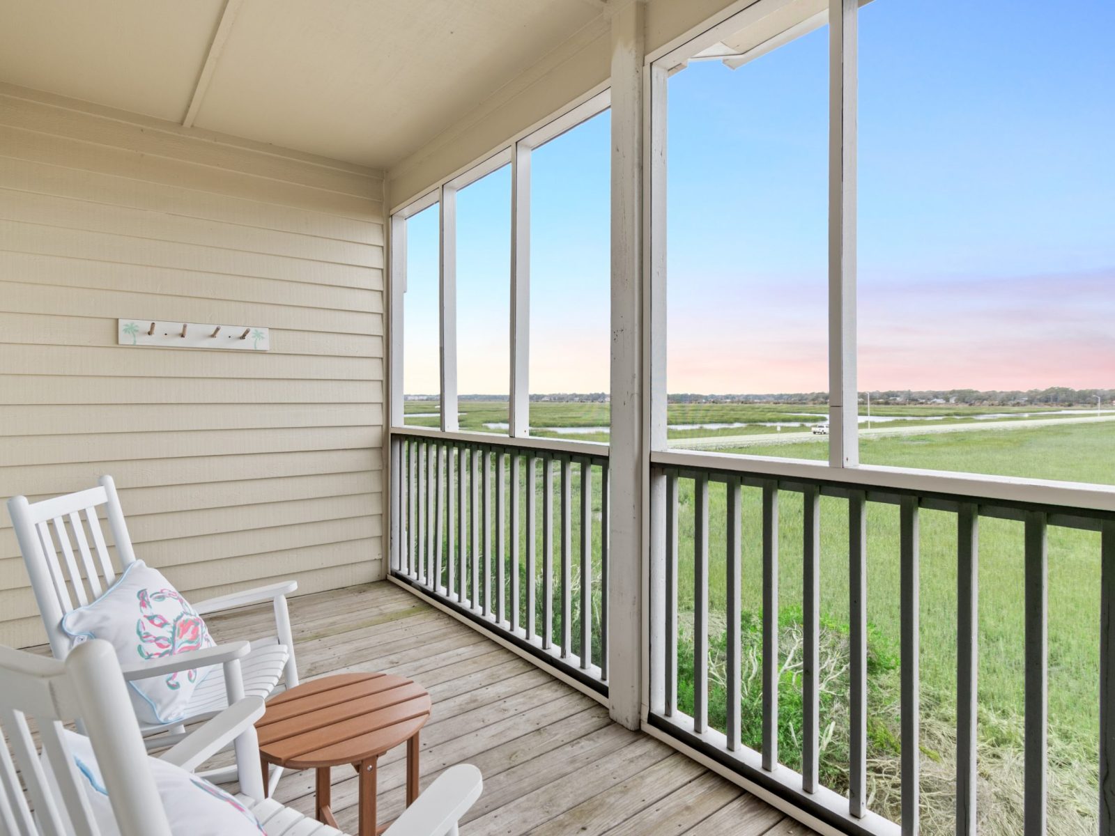 Screened porch with two white rocking chairs and a small round table overlooks a scenic grassy marshland under a blue and pink sunset sky. Enjoy the view from your Sunset Inn top floor room, with wooden flooring and beige siding.