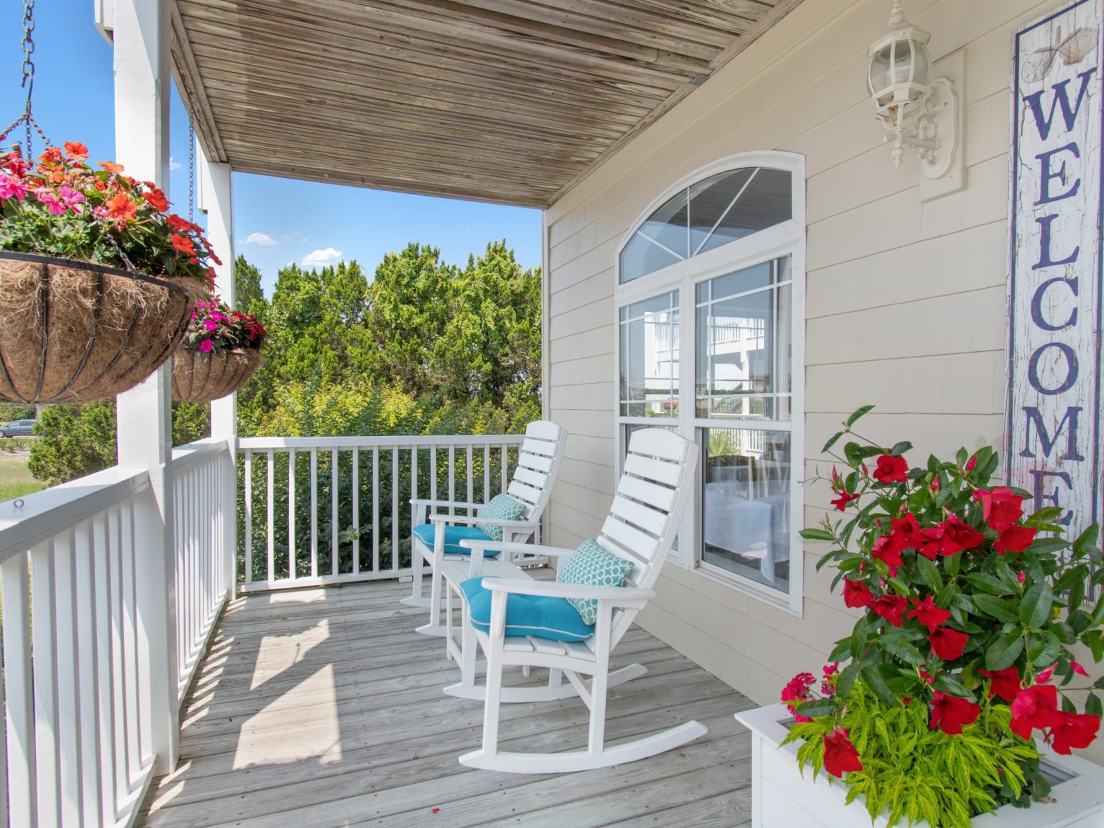 A cozy porch with two white rocking chairs, blue cushions, potted red flowers, hanging flower baskets, a “Welcome” sign, and white railings overlooking greenery—perfect for relaxing after exploring Sunset Inn special offers.